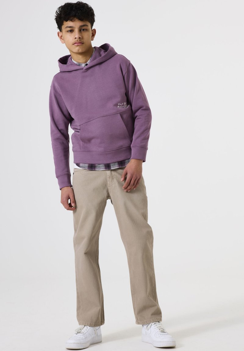 Un chico adolescente de pie, vestido con una sudadera morada, pantalones beige, zapatillas blancas y una camisa de capas debajo, contra un fondo blanco liso.