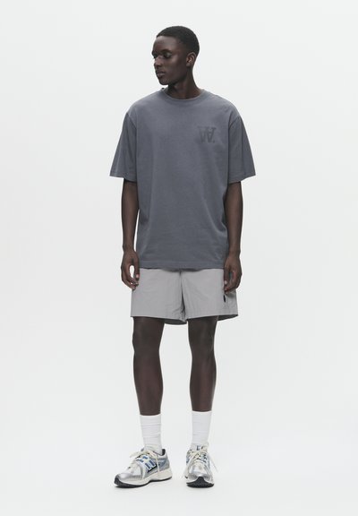 Grå oversized bomulds t-shirt med en logo på venstre bryst, parret med lysegrå shorts og sølvfarvede løbesko, hvide sokker.