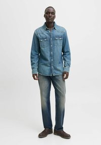 Cămașă din denim de culoare albastru deschis, cu două buzunare pe piept, închidere cu nasturi și cusături contrastante; asortată cu blugi desenați în albastru și pantofi maro.