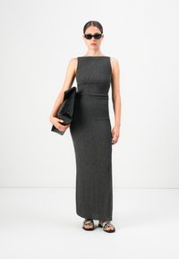 Ženska v teksturirani brezrokavni temnosivi maksi obleki, drži velik črn clutch, nosi črne sončna očala in sandale s kačjim vzorcem.