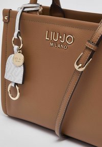 Sac à main en cuir marron texturé avec logo doré, bandoulière réglable et étiquette d'accent détachable blanche et grise avec un fermoir circulaire.