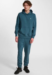 Felpa teal con tasca a marsupio e polsini a costine, abbinata a pantaloni sportivi coordinati con polsini elasticizzati. Scarpe da ginnastica nere.