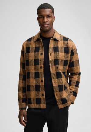 Bruin en zwart geblokt corduroy jasje met een kraag, knoopsluiting en borstzakken, gedragen over een zwart shirt.