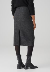 Femme portant une jupe midi en maille côtelée gris foncé avec une fente à l'arrière, des collants noirs, des bottines noires et un haut noir à manches longues, debout sur un sol gris.