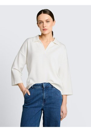 Femme aux cheveux foncés attachés, portant un haut blanc à manches longues avec col et un jean bleu taille haute, debout avec une main dans la poche.