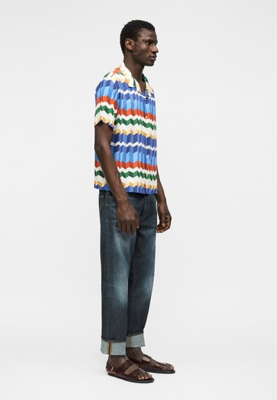 Άνδρας που φοράει πολύχρωμο πουκάμισο με ζικ-ζακ μοτίβο, σκουρόχρωμα τζιν με γυρισμένα μπατζάκια και καφέ σανδάλια με ανοιχτά δάχτυλα, σε έναν μονόχρωμο φόντο.