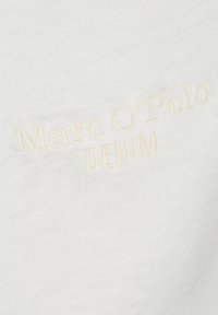 Witte stof met een zachte textuur heeft geborduurde "Marc O'Polo" en "DENIM" in lichtbeige. Close-up detail toont de kwaliteit van de stiksels.
