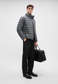 Pelēks pūkains mētelis ar augstu apkakli, horizontālu quilted rakstu un priekšējo rāvējslēdzēju, saslēgts ar tumšiem biksēm un melnu somu.