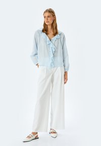 Lichtblauwe blouse met gerimpelde voorkant, knoopsluiting, lange mouwen. Gecombineerd met witte wijde broek en witte puntige schoenen met accenten.