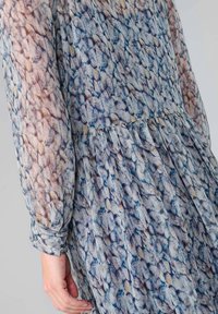 Un vestido transparente de manga larga presenta un patrón texturizado en azul y beige con sutiles pliegues en la cintura y puños abotonados.