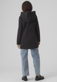 Chaqueta negra con capucha y acabado mate liso, combinada con vaqueros azul claro de corte amplio y zapatos planos negros. Diseño minimalista, sin patrones visibles.