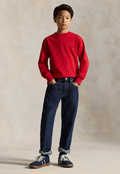Boy stands with hands in pockets wearing red long-sleeve shirt, dark blue rolled-up jeans, and navy sneakers against neutral background.