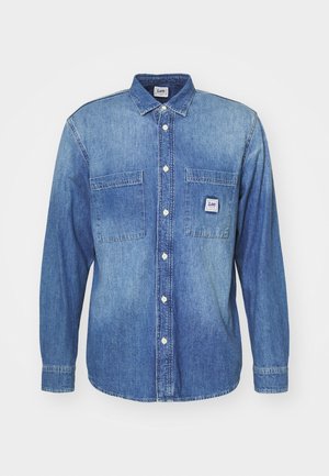 Jeansowa koszula z długim rękawem w jasnoniebieskim kolorze, z dwoma kieszeniami na piersi, zapięciem na guziki z przodu oraz metką z logo na lewej kieszeni.