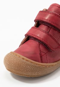 Chaussure rouge en cuir avec deux bandes Velcro, bout arrondi et semelle en caoutchouc texturée. Présente un design cousu et une finition mate.