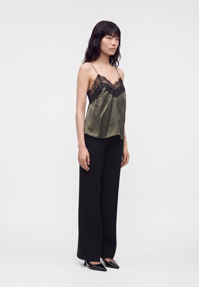 Femme aux longs cheveux noirs portant un caraco en satin vert avec de la dentelle noire et un pantalon large noir, debout devant un fond uni.