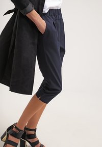 Žena v zkrácených námořnických kalhotách s elastickým pasem, černých sandálech na vysokém podpatku, bílém topu a černém dlouhém sakě, ruka v kapse.