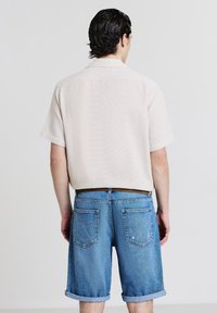 Uomo con capelli scuri che indossa una camicia a maniche corte beige chiaro con texture e pantaloni corti in denim blu arrotolati, in piedi di fronte a un muro bianco semplice.