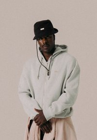 Grauer Zip-Hoodie mit Kapuze, ausgestattet mit Kordeln und einem goldenen Logo. Schwarzer Bucket Hat aus strukturiertem Material mit verstellbarem Riemen.