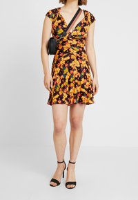 Kwiatowa sukienka w stylu wrap z żywymi pomarańczowymi i żółtymi kwiatami na czarnym tle, z krótkimi rękawami i rozkloszowanym dołem. Połączona z czarnymi szpilkami.