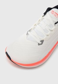 Λευκό αθλητικό παπούτσι με επάνω μέρος από πλέγμα, με υφή, μαύρες λεπτομέρειες στη γλώσσα και φωτεινή πορτοκαλί εξωτερική σόλα.