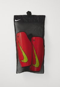 Punased Nike säärekotid kollaste aksentidega, hoiustatud mustas võrgukotis, mille ülemises osas on käepide mugavaks kandmiseks. Kotil on Nike logo.