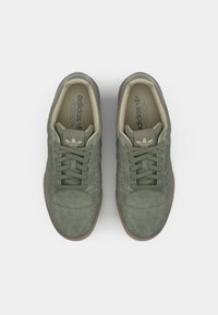 Sapatilhas verdes em camurça com biqueira redonda, atacadores, detalhes perfurados e forro bege. Apresenta o logo da Adidas na língua e nas laterais.