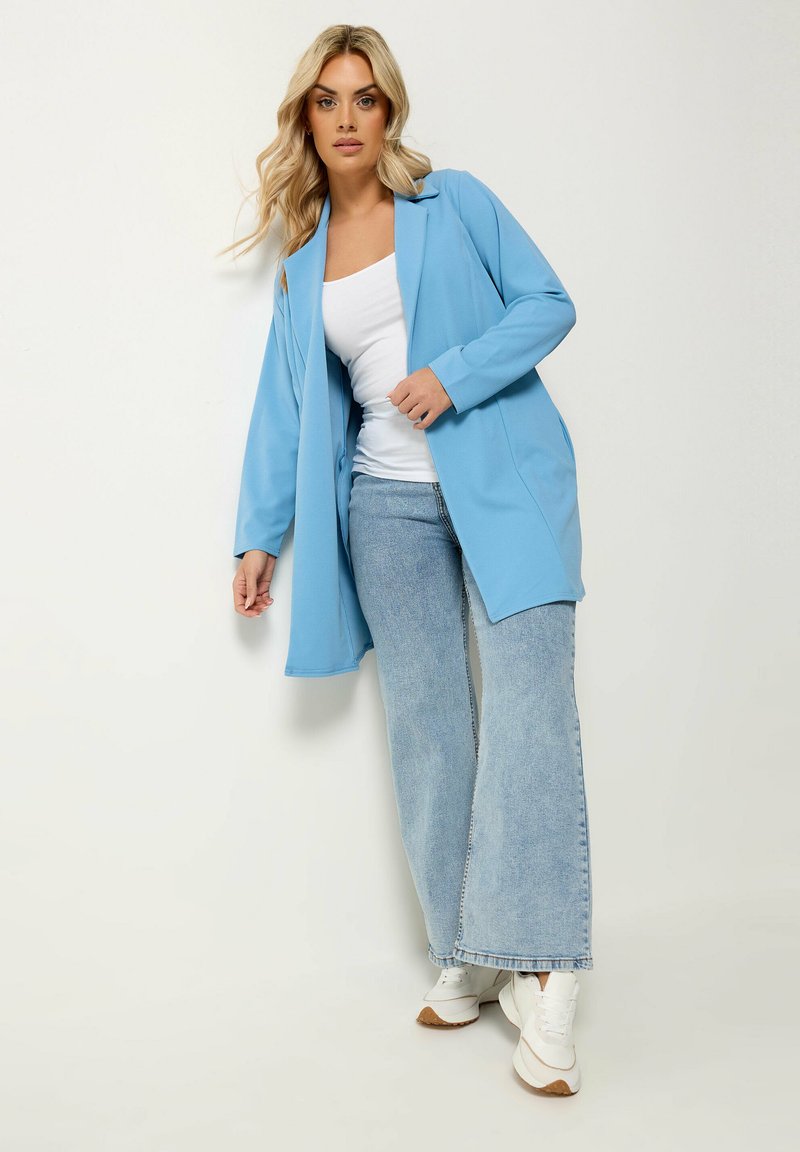 Vrouw die een lichtblauwe blazer, wit tanktop, jeans met wijde pijpen en witte sneakers draagt tegen een effen witte achtergrond.