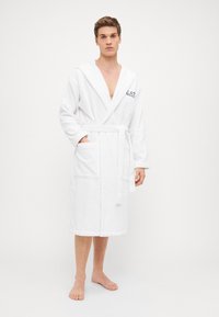 Accappatoio bianco in morbido tessuto terry, con maniche lunghe, cappuccio, cintura e due tasche frontali. Il design è semplice e funzionale.