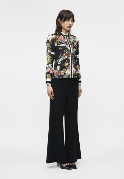 Γυναίκα που φοράει μαύρο μπλουζάκι με λουλούδια και λευκή διακόσμηση, καθώς και φαρδιά μαύρη παντελόνα, στέκεται και κοιτάζει ελαφρώς προς τα δεξιά με φόντο λευκό.