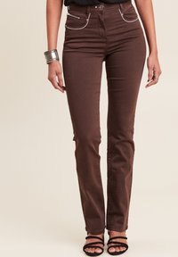 Braune Denim-Jeans mit schlanker Passform, versehen mit weißen Kontrastnähten, Fronttaschen und einem Reißverschlusdetail. Kombiniert mit schwarzen Sandalen mit Riemen.