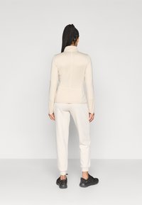 Ζακέτα με φερμουάρ σε κρεμ χρώμα με μακριά μανίκια, συνδυασμένη με παντελόνι σε ανοιχτό χρώμα και μαύρα αθλητικά παπούτσια. Λείο ύφασμα, εφαρμοστή γραμμή.