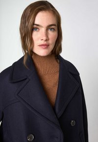 Marineblauwe wollen blend jas met een dubbelbreasted ontwerp, grote revers en een bruine ribgebreide coltrui eronder. Soepele textuur, ronde knopen.