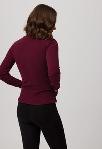 Top de manga larga burdeos con acabado texturizado, diseño ajustado y aberturas laterales, combinado con leggins negros. Vista trasera visible.