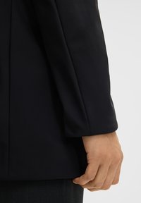 Donker, getailleerde blazersleeve met een zachte textuur, voorzien van een strakke, rechte snit en subtiele stikseldetails. Hand rustend nabij de zoom.