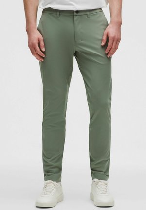 Mann trägt olivgrüne, eng anliegende Hose und weiße Sneakers, steht vor einem schlichten hellen Hintergrund mit entspannt an den Seiten hängenden Händen.