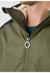 Olijfgroene jas met een ritssluiting aan de voorkant, voorzien van een zachte crèmekleurige voering, zwarte schouderaccenten en een ronde zilveren ritstrekker.