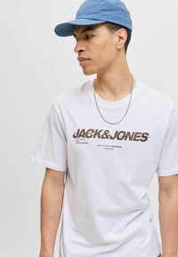 Tricou alb din bumbac cu logo-ul "JACK&JONES" în culoarea maro, mâneci scurte, gât rotund, purtat cu o lanț argintie și o șapcă albastră deschisă.