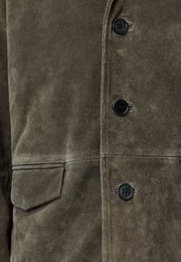 Cappotto in suede verde oliva con una texture morbida, dotato di una tasca frontale e bottoni neri su una chiusura dritta. Dettagli di cucitura sottili visibili.