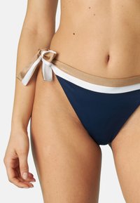 Slip di bikini blu navy con vestibilità bassa, caratterizzato da un bordo beige e bianco in vita annodato a fiocco. Texture liscia, copertura minima.