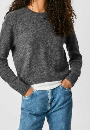 Kvinde med krøllet hår iført en mørk grå, blød sweater over en hvid skjorte, blå jeans, hænderne i lommerne, stående foran en lys baggrund.