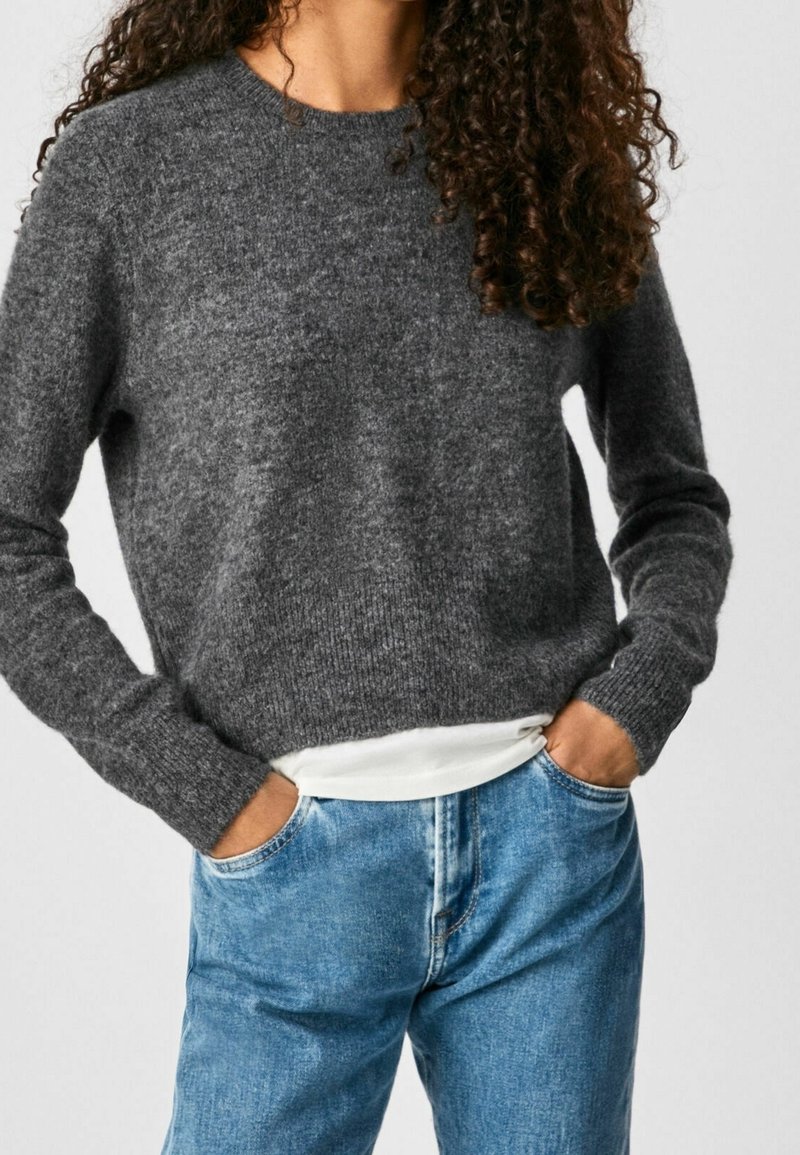 Femme aux cheveux bouclés portant un pull en laine gris foncé par-dessus une chemise blanche, jean bleu, mains dans les poches, debout devant un fond clair.