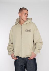 Béžová mikina na zip s předními kapsami, na levém prsu s grafickým designem. Vyrobena z hustého materiálu, má volný střih a kapuci na šňůrku.