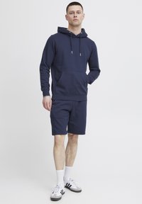 Marineblauwe katoenen hoodie met een voorzak en een trekkoordkap, gecombineerd met bijpassende shorts, weergegeven op een persoon die rechtop staat.