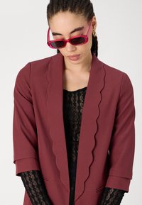 Blazer bordeaux avec des bords en dentelle, tissu doux, manches trois-quarts, associé à un top en dentelle noire et des lunettes de soleil roses.
