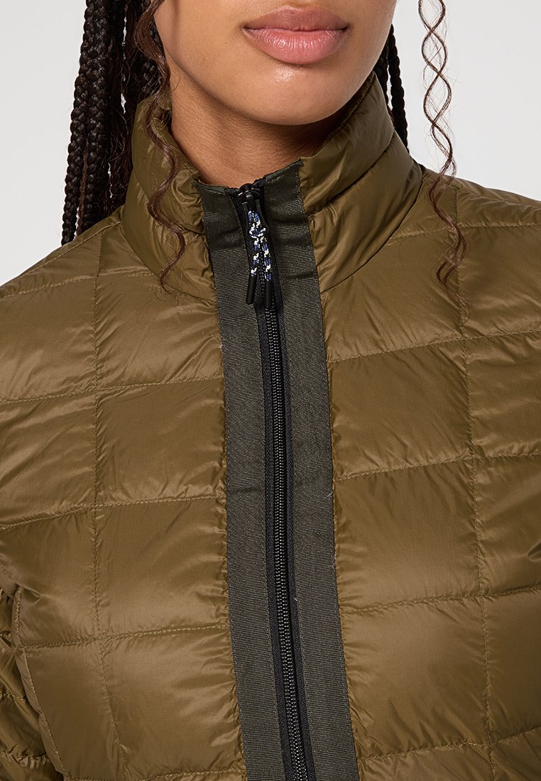 Junge Person mit geflochtenem Haar, die eine olivgrüne gesteppte Jacke trägt, die bis zum Kragen zugezippt ist.