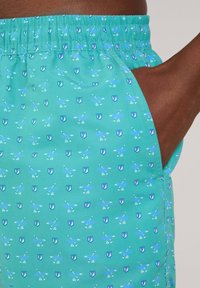 Pantaloni da bagno turchesi con cintura elastica, caratterizzati da un motivo ripetuto di scudi blu e pagaie bianche. Inclusi tasche laterali.