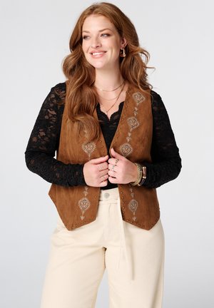 Vrouw met lang rood haar draagt een bruine geborduurde vest over een zwarte kanten top en crèmekleurige broek, glimlachend en opzij kijkend.