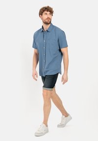 Lyseblå kortærmet skjorte med knapper og brystlomme, parret med mørke denimshorts. Modellen har hvide sneakers på og viser et afslappet look.
