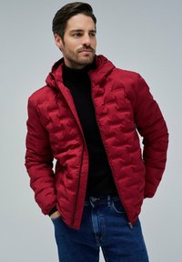 Hombre vistiendo una chaqueta acolchada roja sobre un suéter de cuello alto negro y jeans azules, con las manos en los bolsillos, mirando ligeramente hacia un lado.