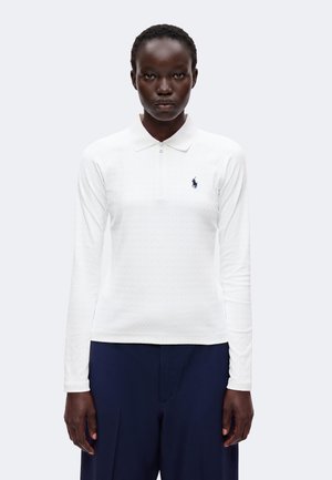 Femme portant un polo blanc à manches longues avec un logo bleu marine et un pantalon bleu marine, debout devant un fond clair uni.